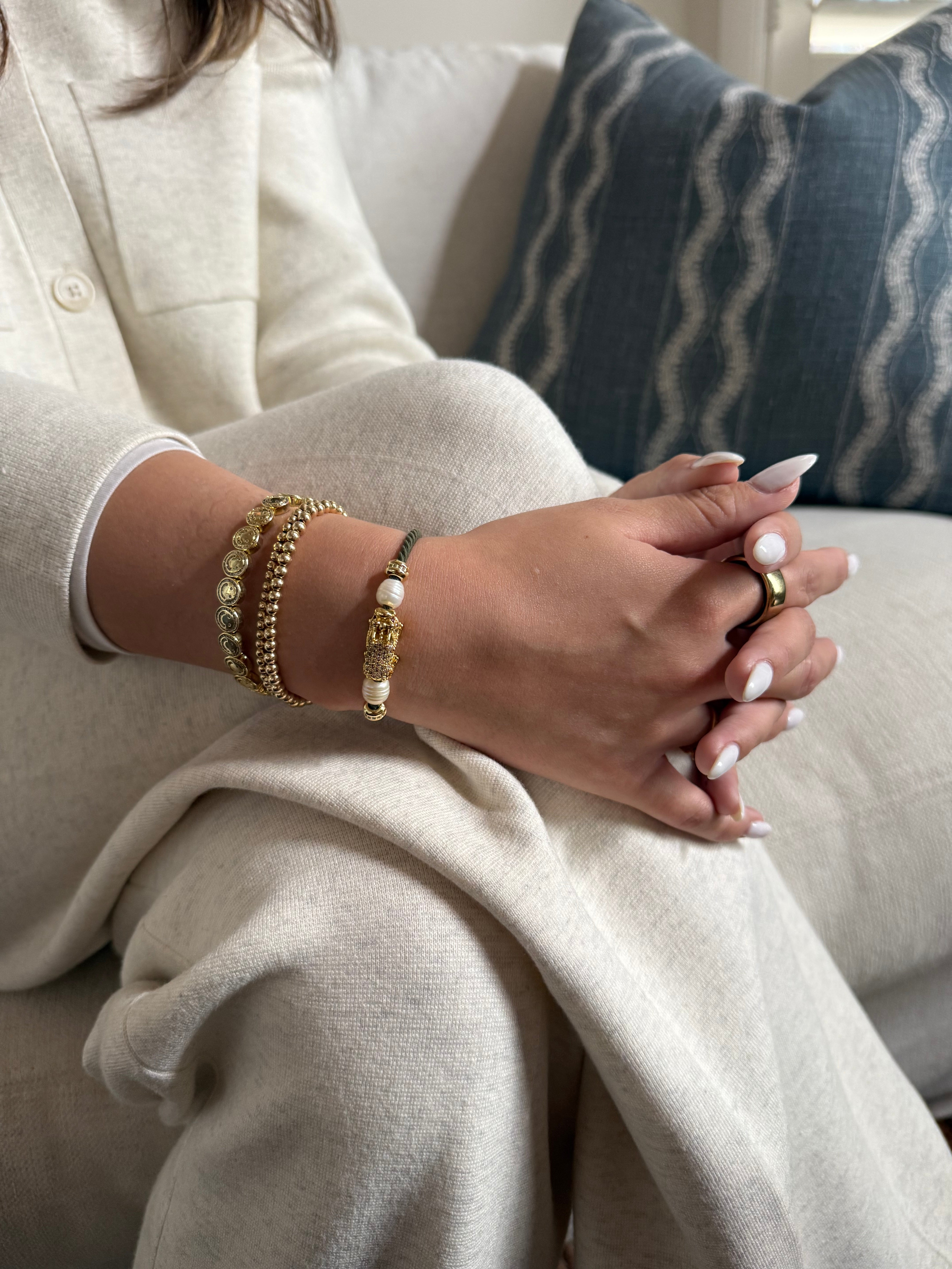 Close-up of a person's hands with gold plated and CZ dragon charm, pearls on green cord hair tie, gold bracelets and a ring, wearing a light-colored sweater.
