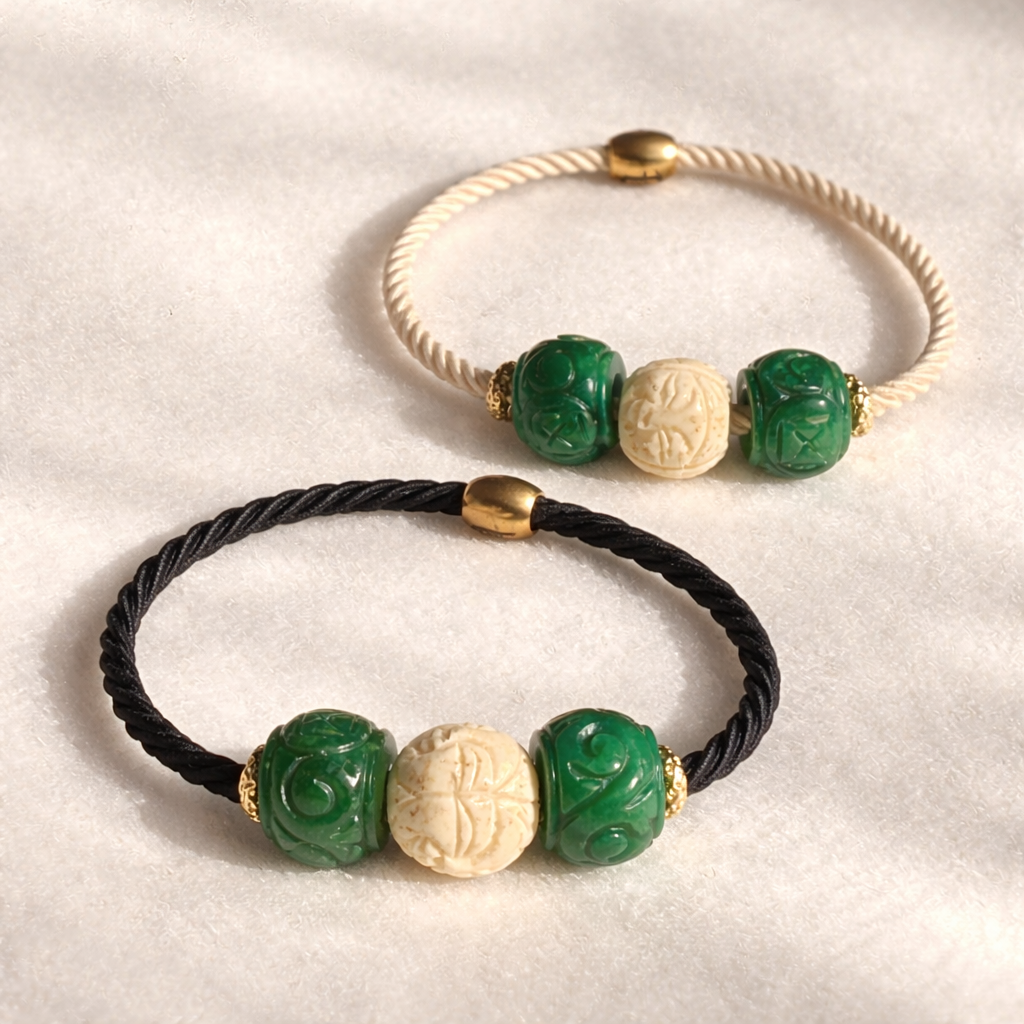 Two hair ties, one black and one beige cord, with green, hand carved ivory bead featuring face, and gold beads on a light background.