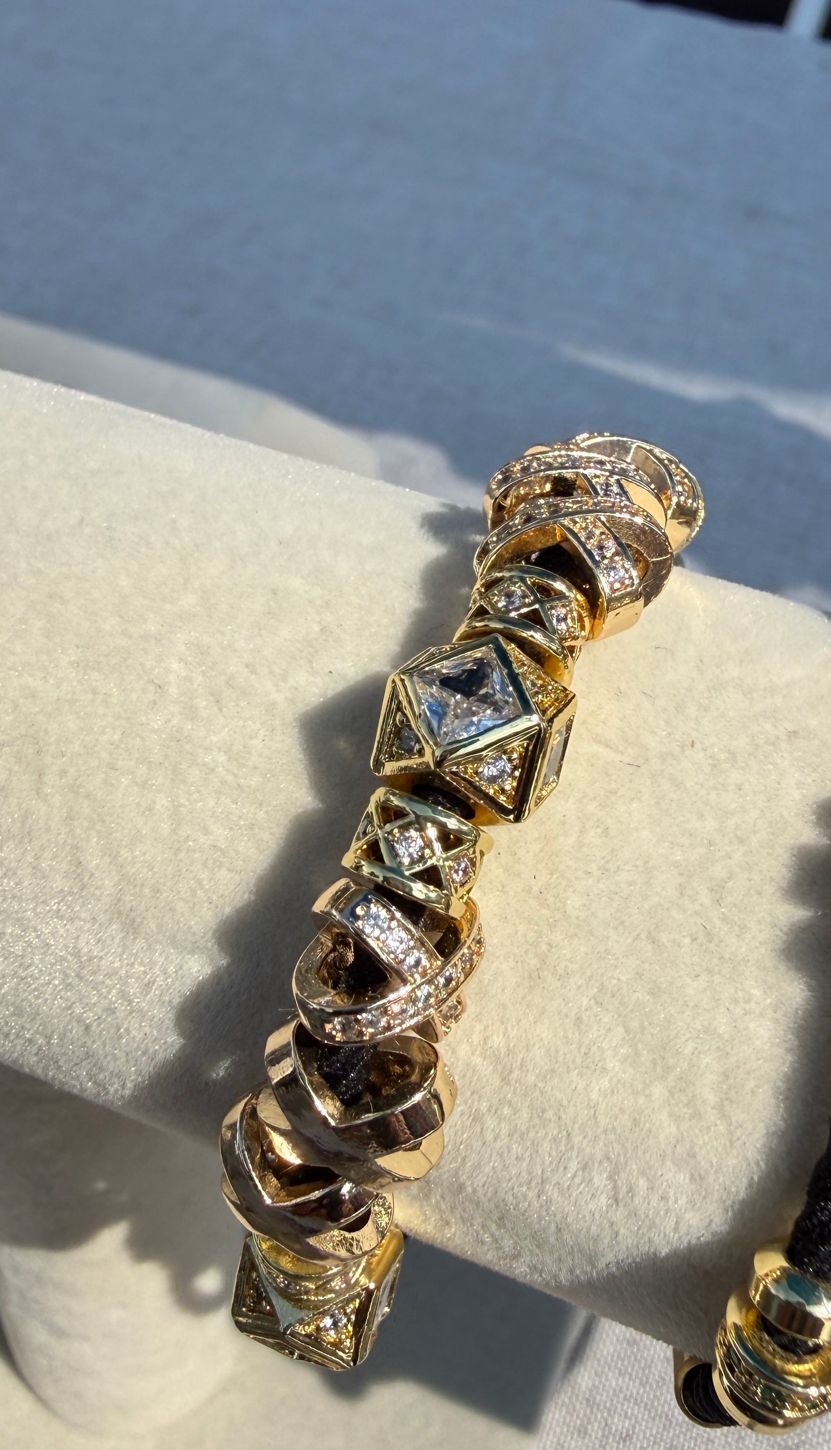 A yellow gold-plated multi-charmed bracelet with geometric cube and spiral links, embellished with clear pavé stones, displayed on a cream-colored jewelry stand. Bracelet hair tie. 
