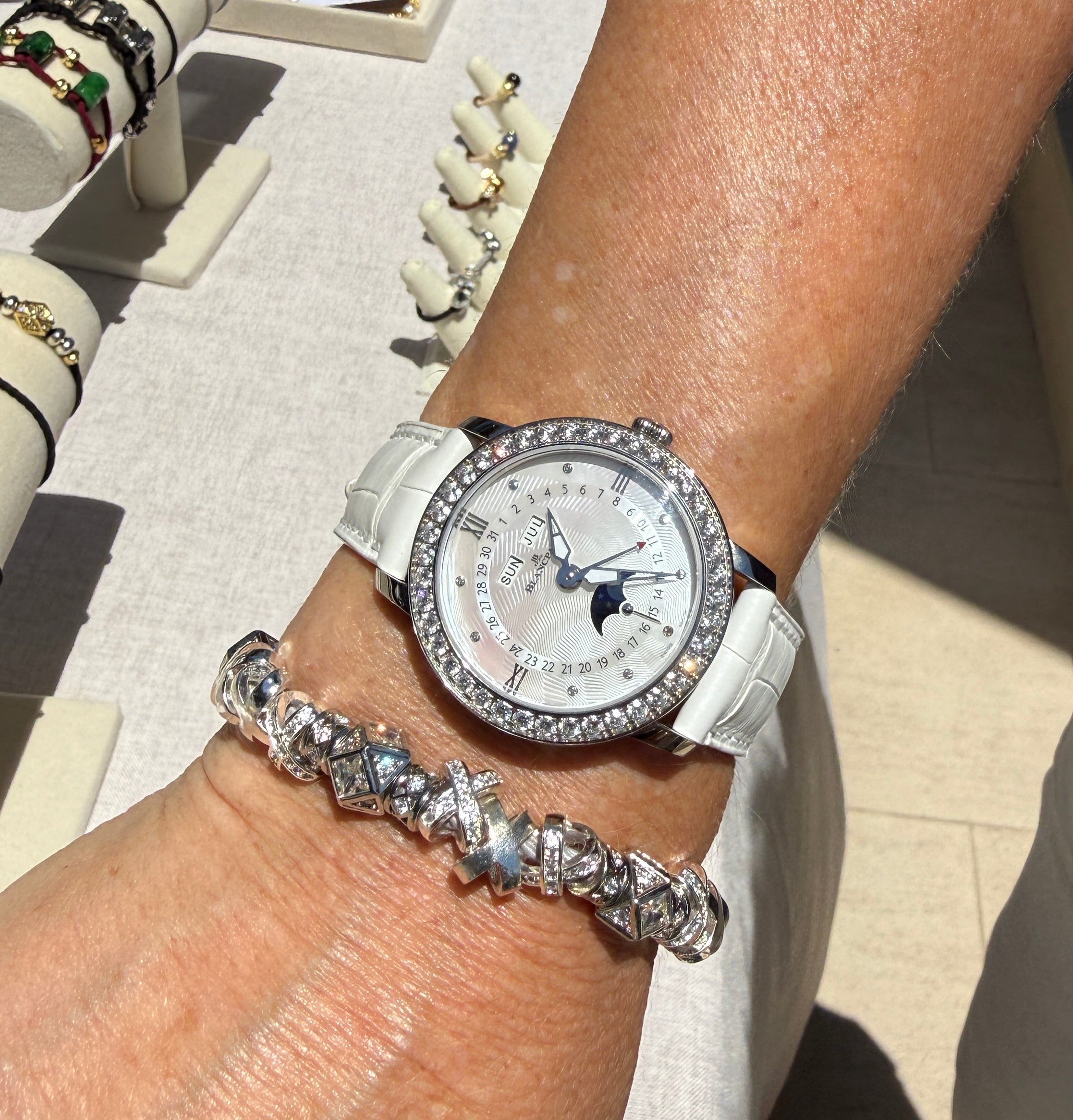 A close-up of a person's wrist wearing a white leather strap watch with a crystal-encrusted silver bezel and a white dial, alongside a chunky platinum plated silver bracelet hair tie with CZs.